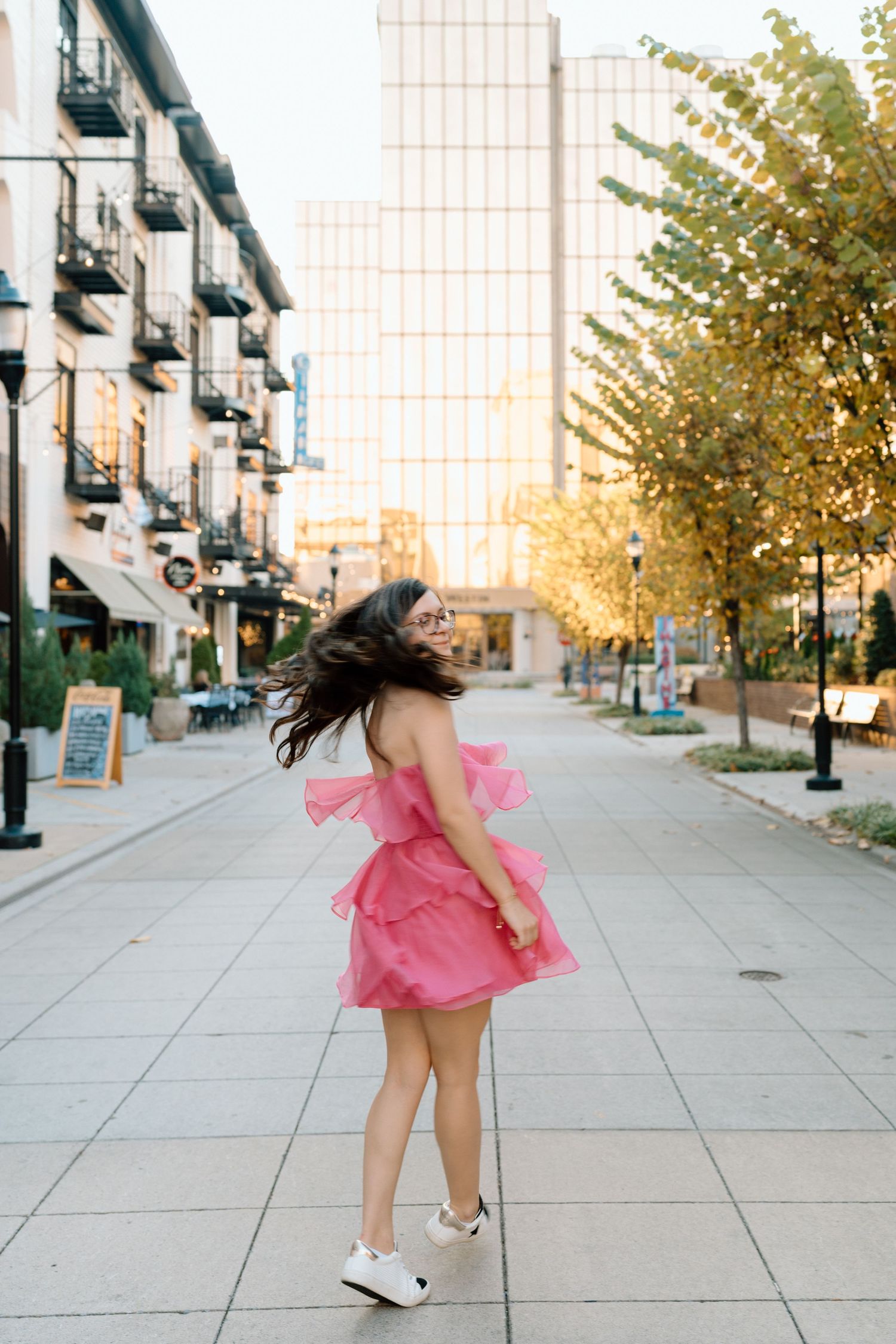 Someone in a pink dress twirls and dances on a sunlit urban sidewalk with modern buildings in the background. Chattanooga Senior Photographe
