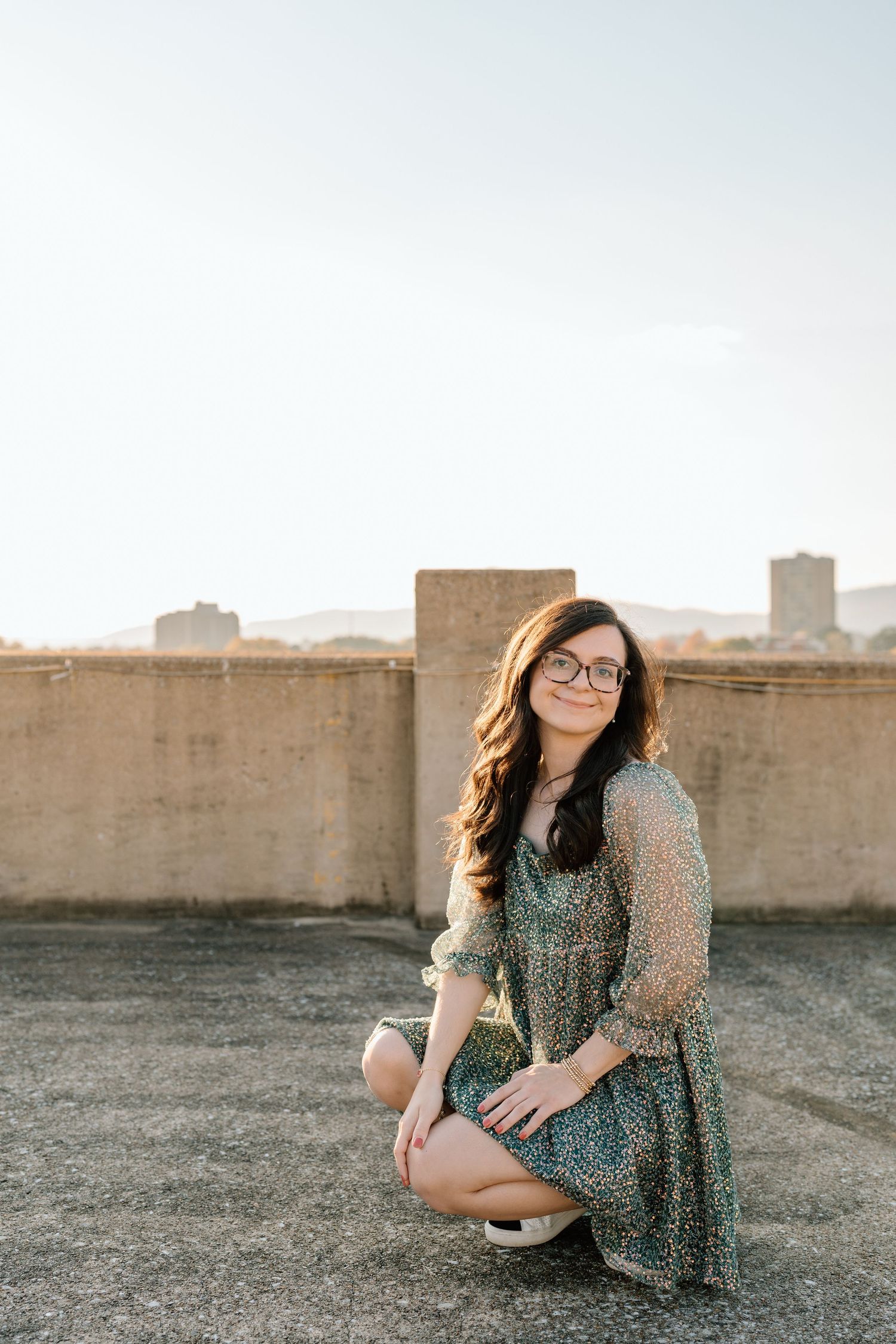A woman in glasses sits casually on a rooftop during sunset with urban skyline behind her. Chattanooga Senior Photographer