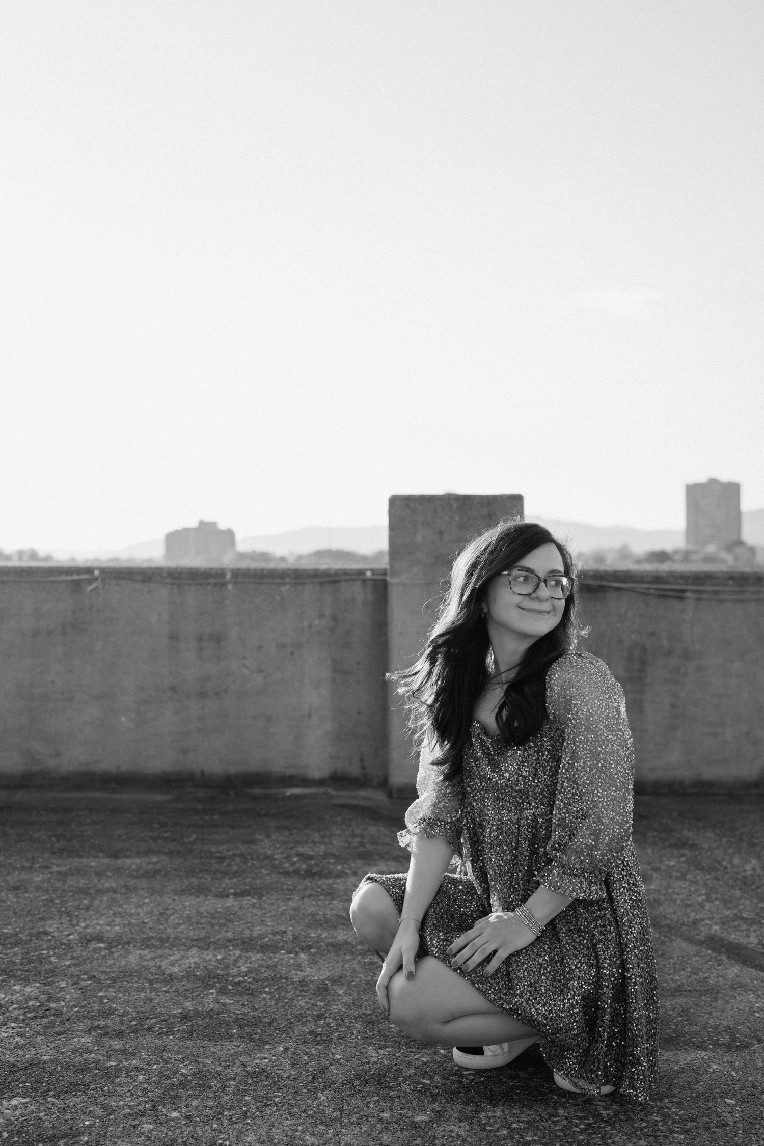 Black and white portrait of person in glasses sitting on rooftop with urban skyline in background. Chattanooga Senior Photographer