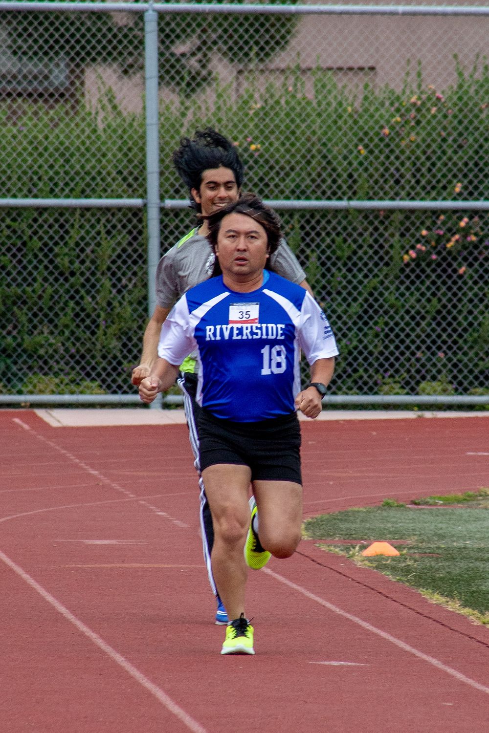 Track athlete in blue Riverside jersey number 18 runs on red track during competition.