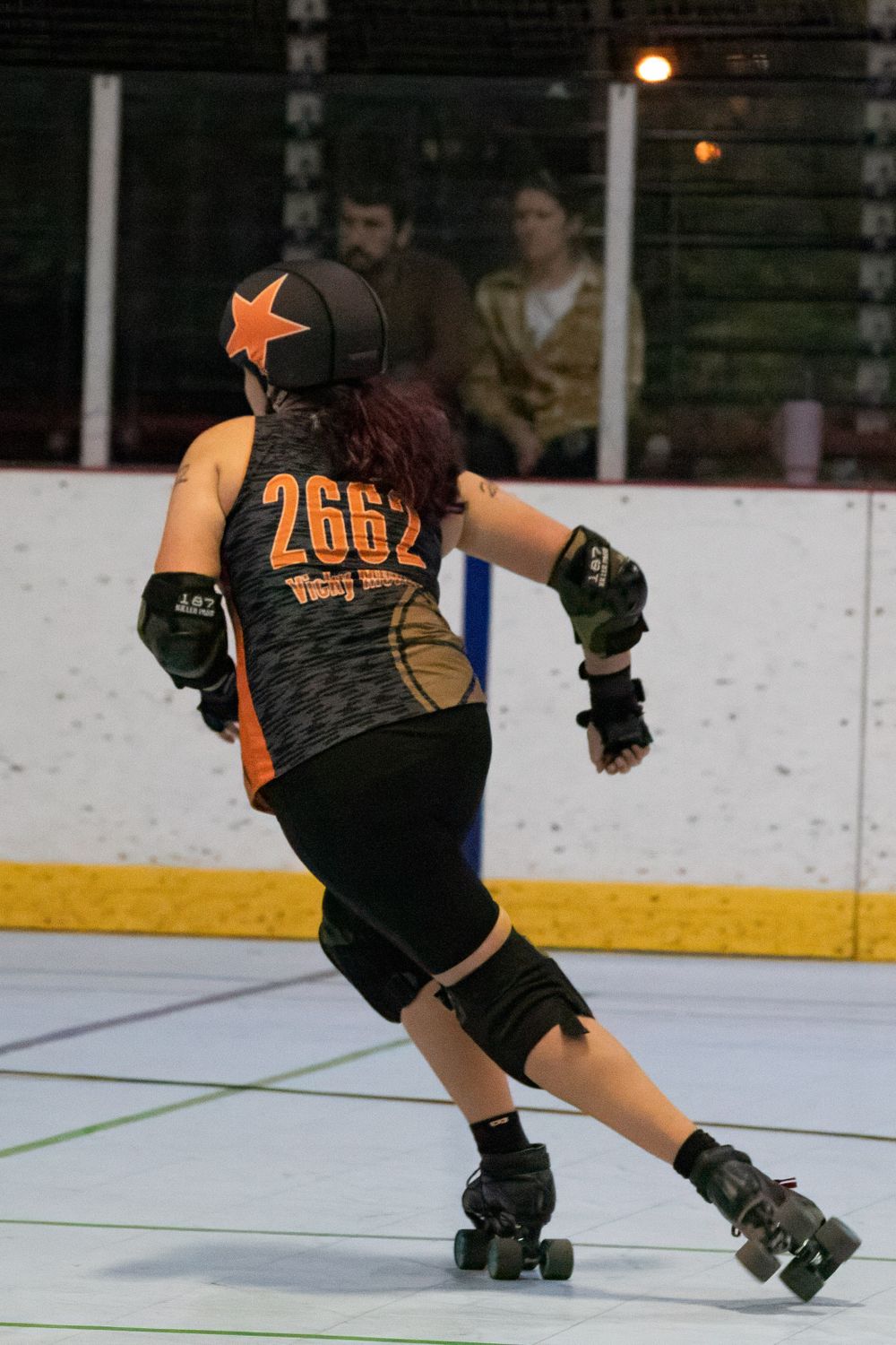 Roller derby skater in orange and black uniform maneuvers on indoor rink.