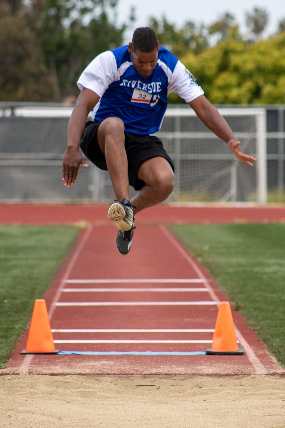 Athlete in blue and white uniform performs dynamic long jump between orange cones on track.