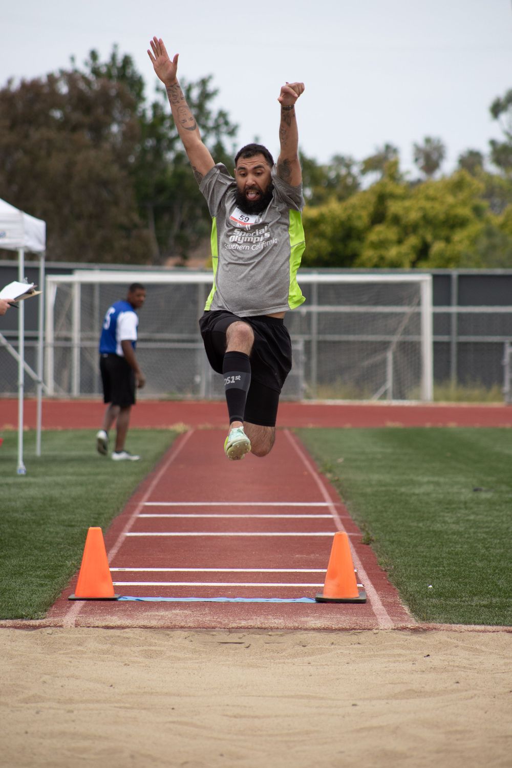 Athlete in gray shirt leaps energetically during long jump competition on outdoor track.
