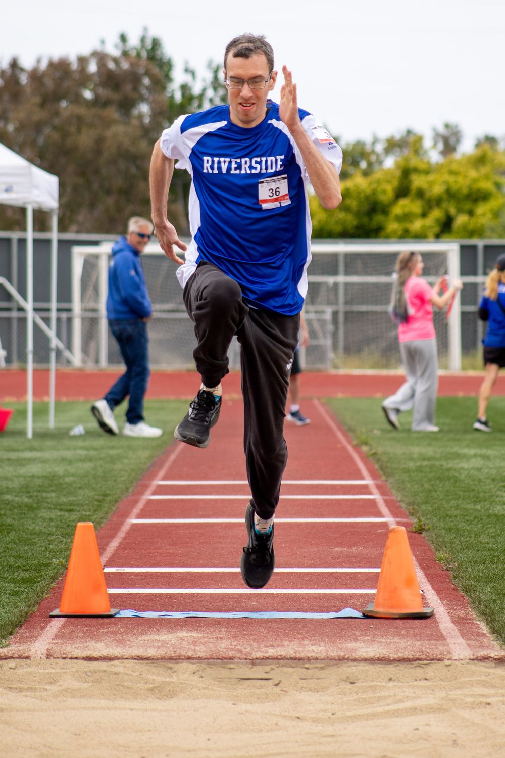 Athlete in blue jersey performs running jump between orange cones on track.