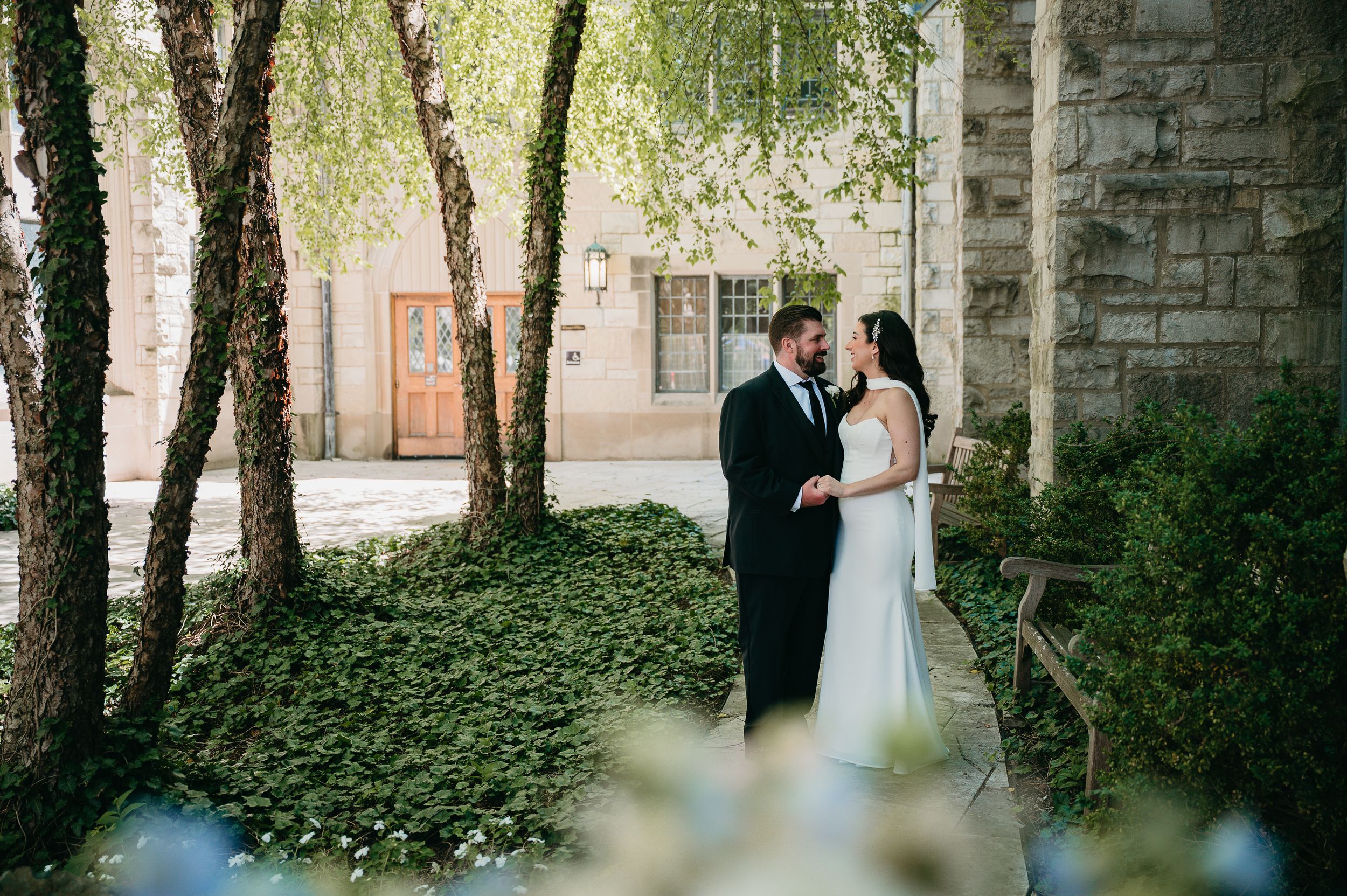 A bride and groom share a romantic moment outdoors among lush greenery and birch trees beside a stone building.