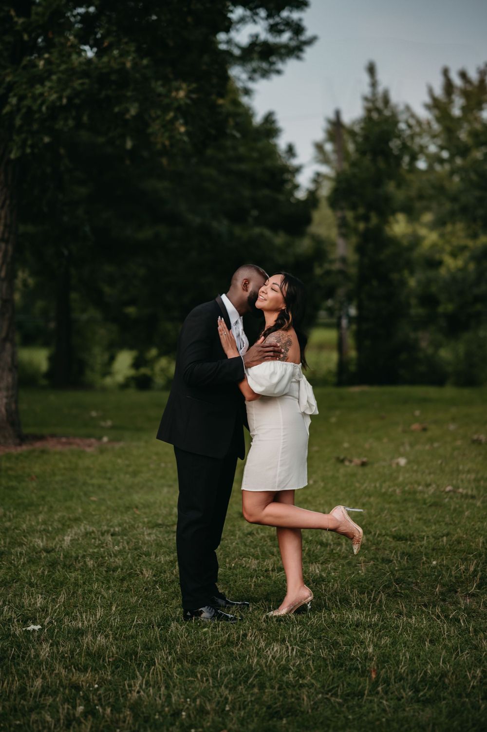 A romantic couple shares a tender moment on a grassy field with trees in the background as the sun sets.
