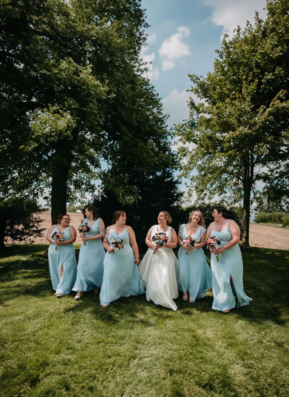 Wedding party in light blue dresses posing together on sunny lawn.