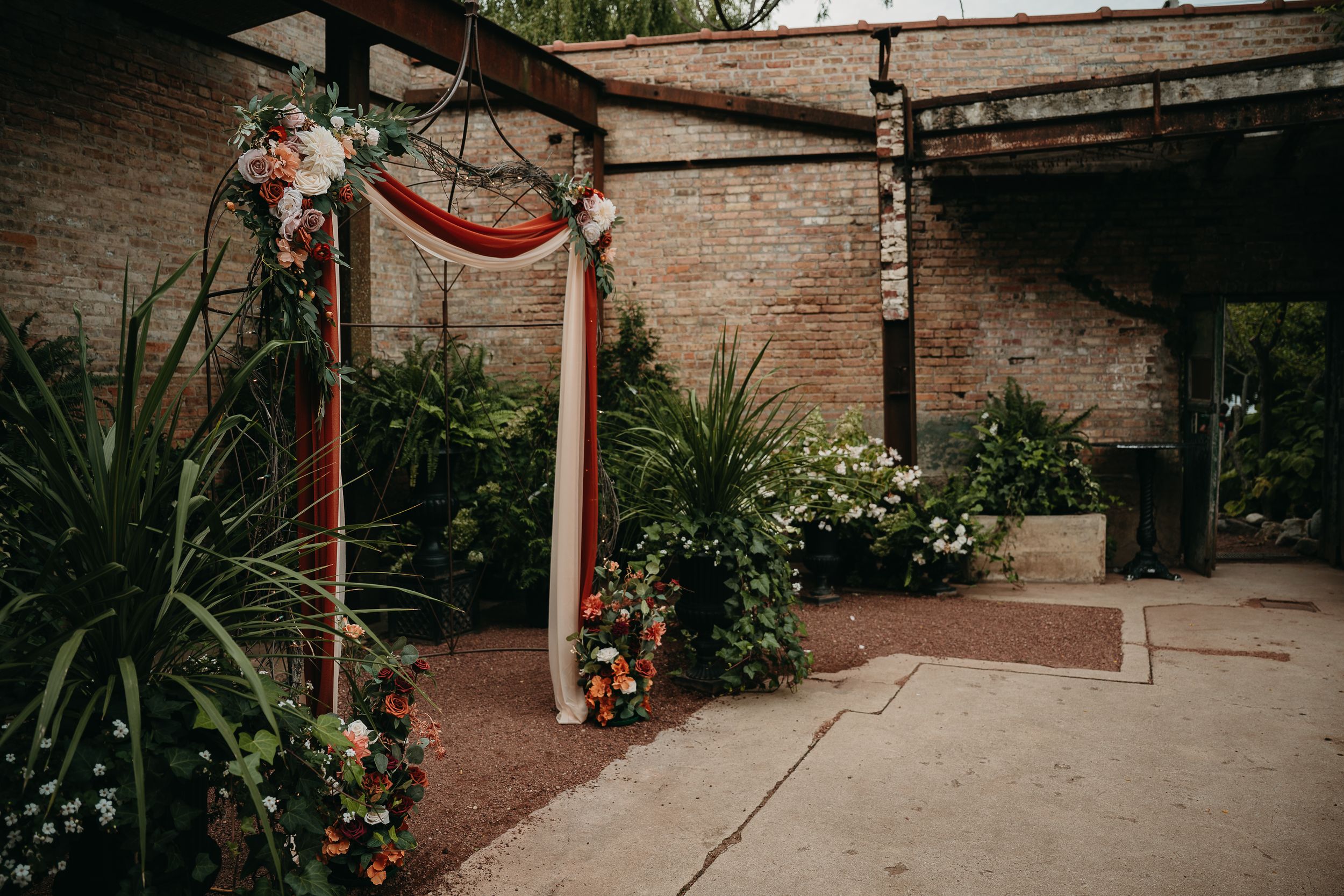 Rustic wedding arch decorated with burgundy fabric and florals stands in a garden courtyard with stone walls and greenery.