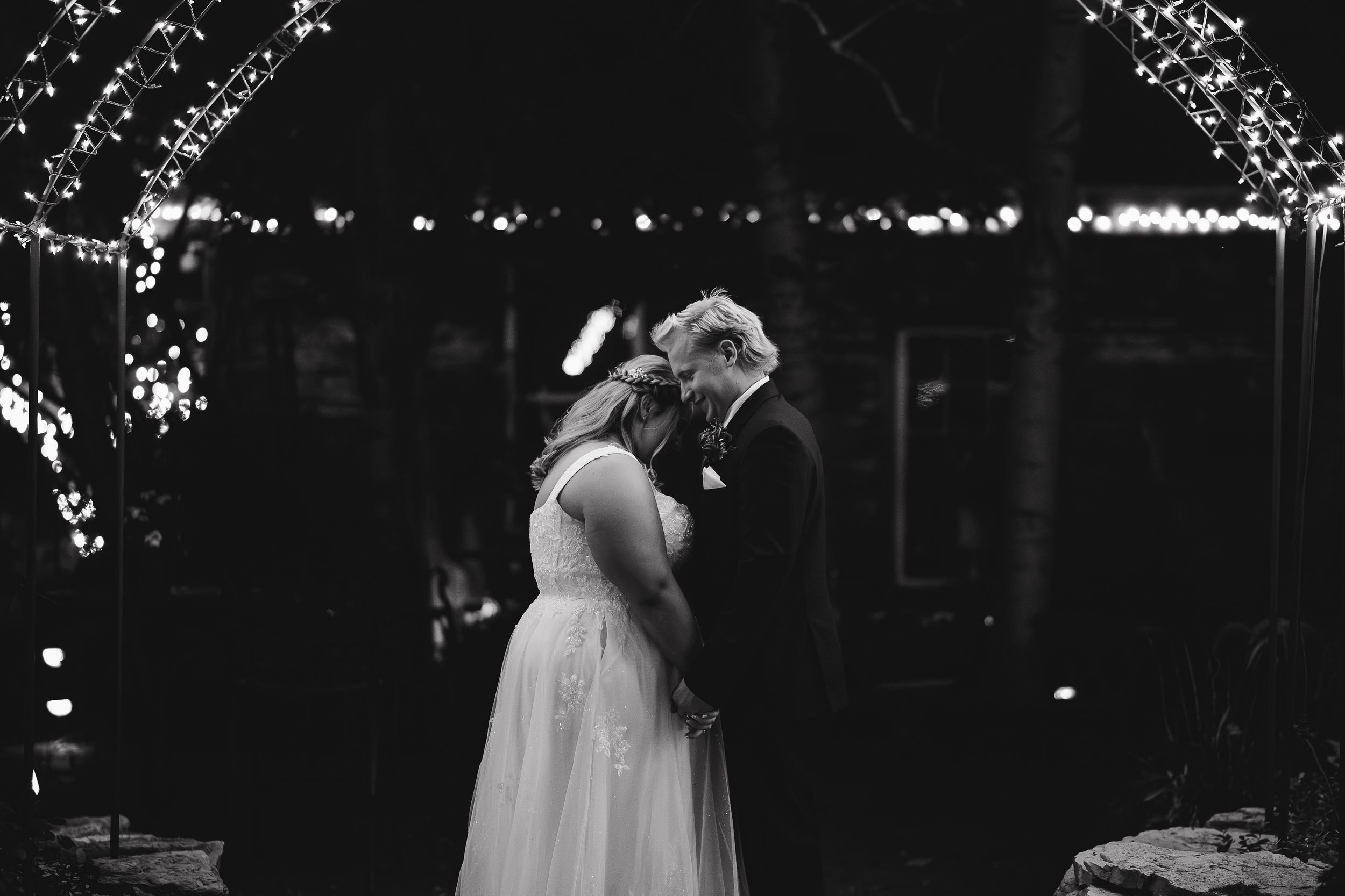 A bride and groom share their first dance on a dark dance floor illuminated by string lights overhead.