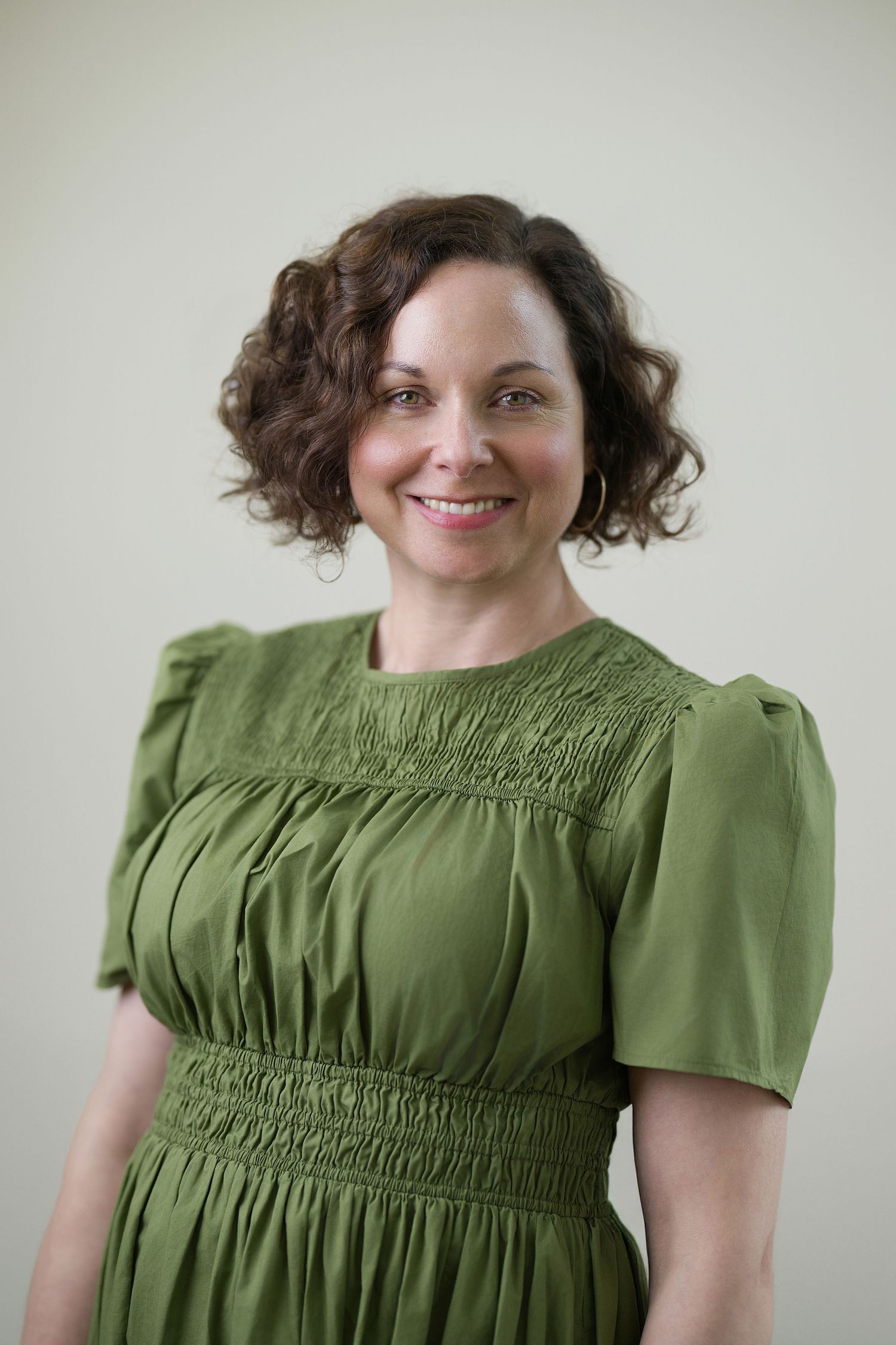 Woman poses in a vintage-style olive green dress with smocked details and puff sleeves–Against cream wall. Professional work photoshoot.
