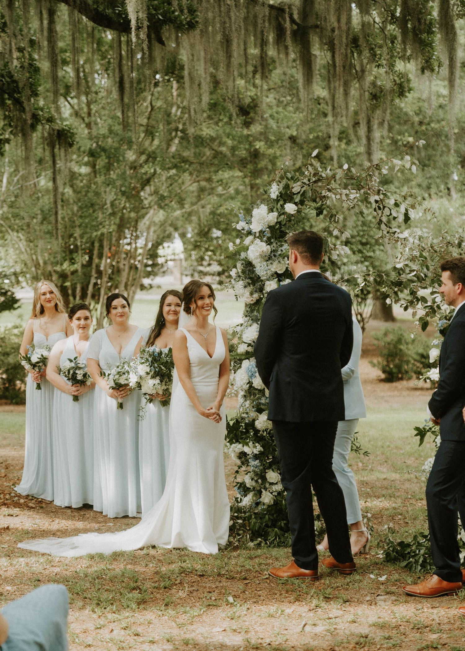A romantic outdoor wedding ceremony under Spanish moss-draped trees with a floral arch and bridesmaids in light blue dresses.