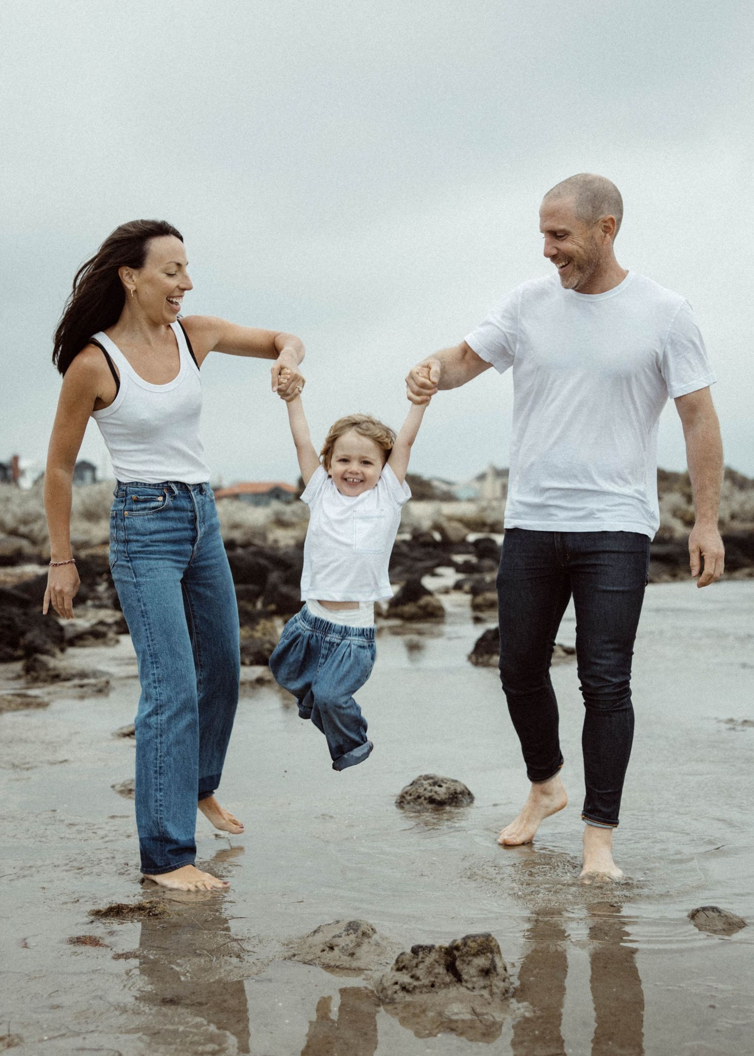 Family in white outfits poses together on dark rocky beach terrain against blue sky and ocean background.