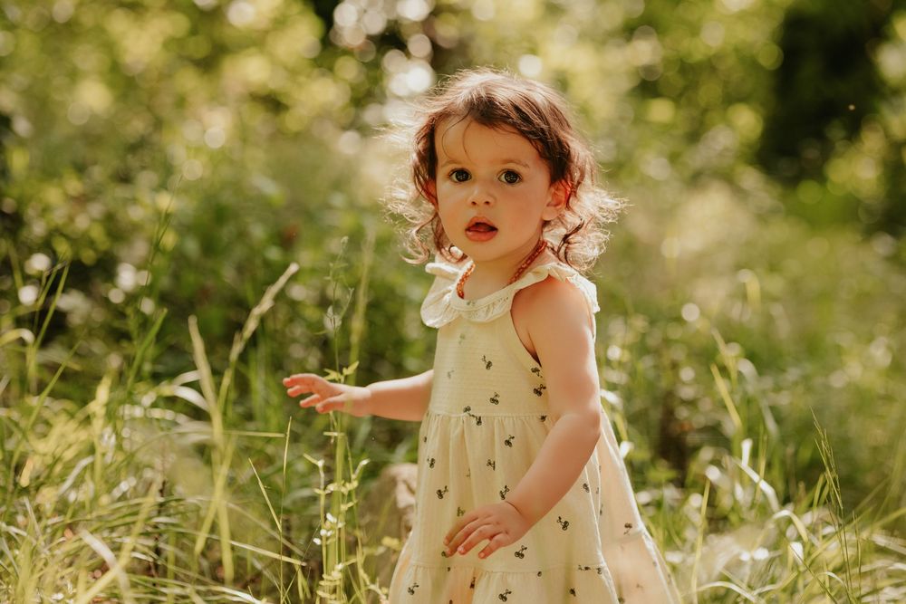 A toddler girl in a floral sundress stands in a lush green meadow, looking back with curious eyes.