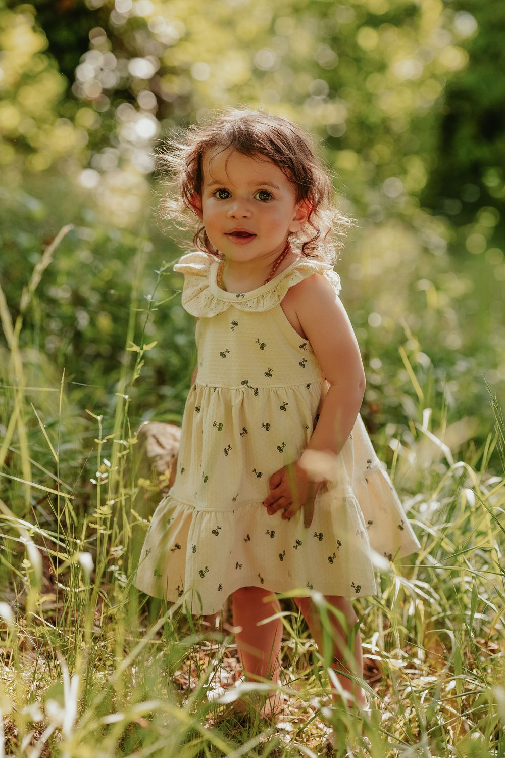 Toddler girl in yellow floral dress standing in tall green grass outdoors in warm sunlight.