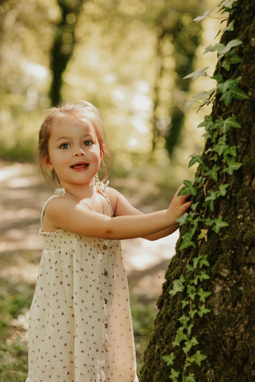 A young girl in a floral dress touches an ivy-covered tree trunk in a sunlit forest, smiling joyfully.