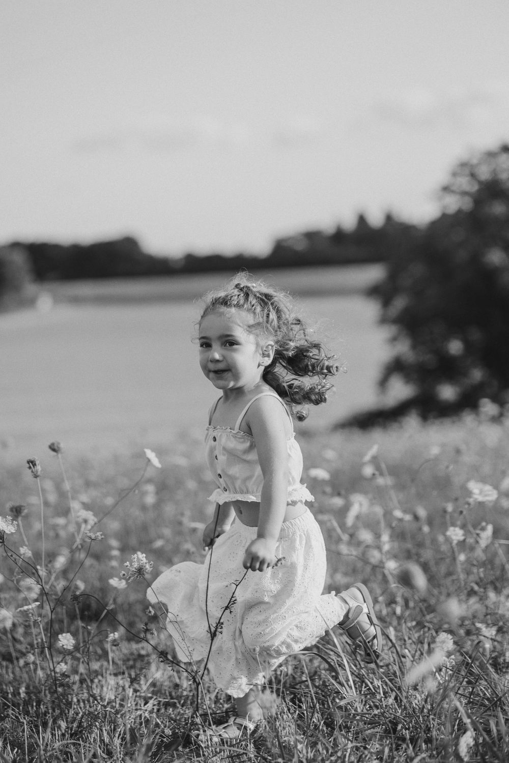 A child in a white dress sits in a field of wildflowers while wind blows through their hair.