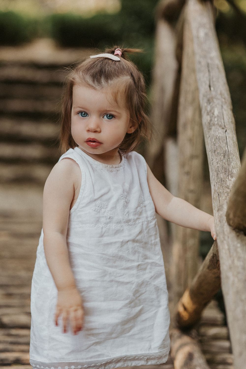 A small child in a white sleeveless dress stands near wooden fencing in an outdoor setting.