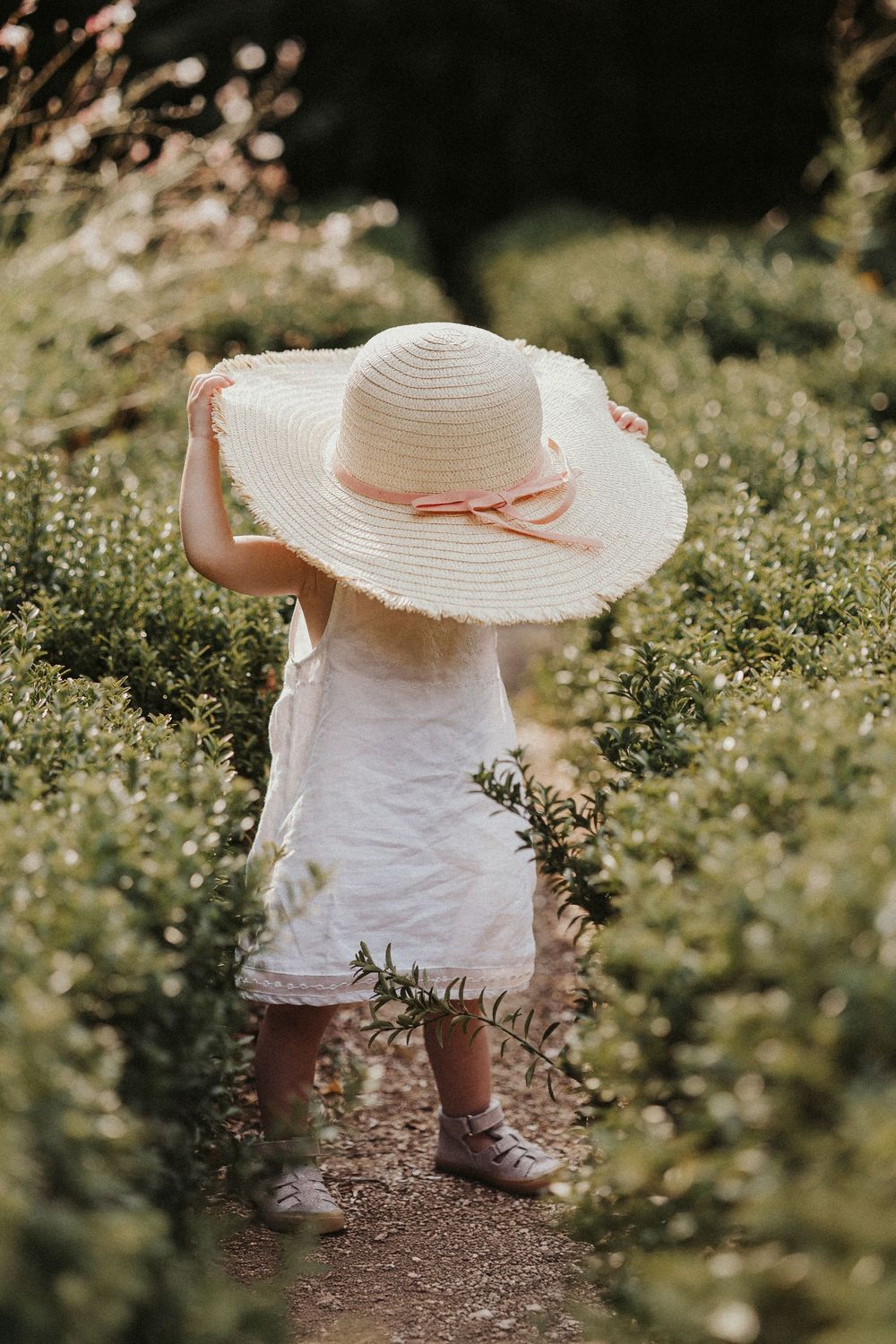 Small feet in brown shoes stand among wildflowers wearing a white dress and sun hat.