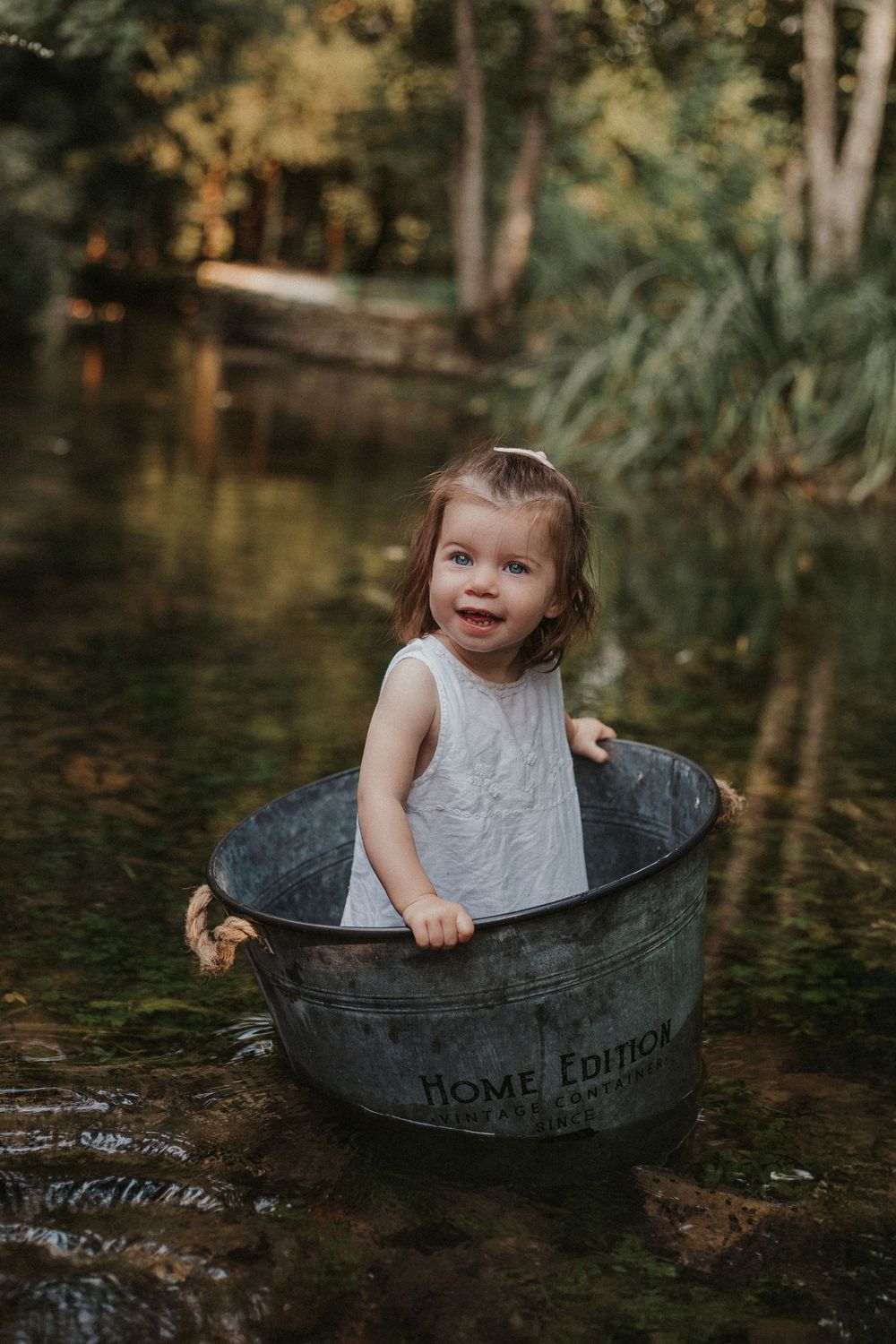 A child sits playfully in a vintage metal washtub surrounded by water and greenery in an outdoor setting.