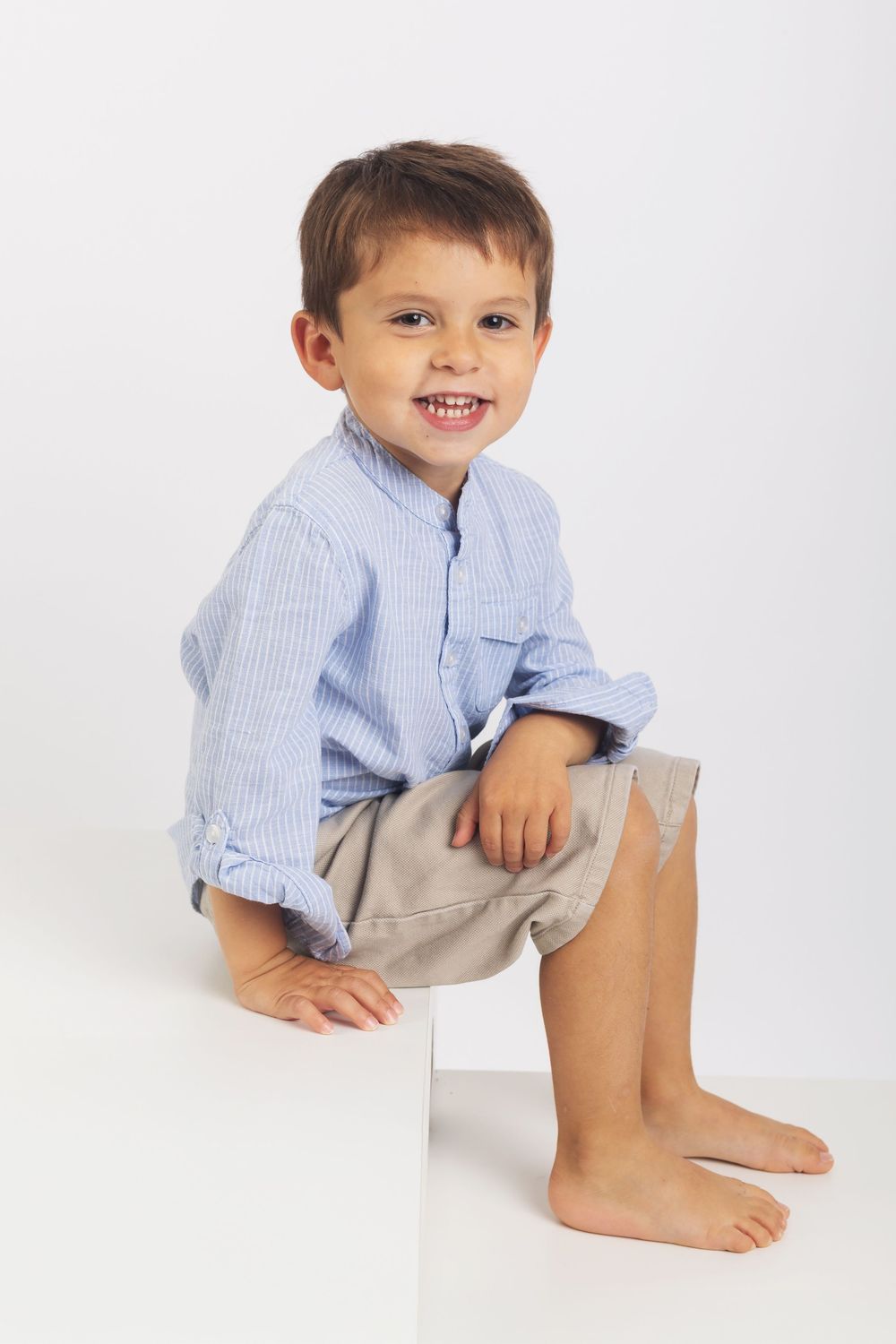 A child in a blue striped shirt and khaki shorts sits on a white background.