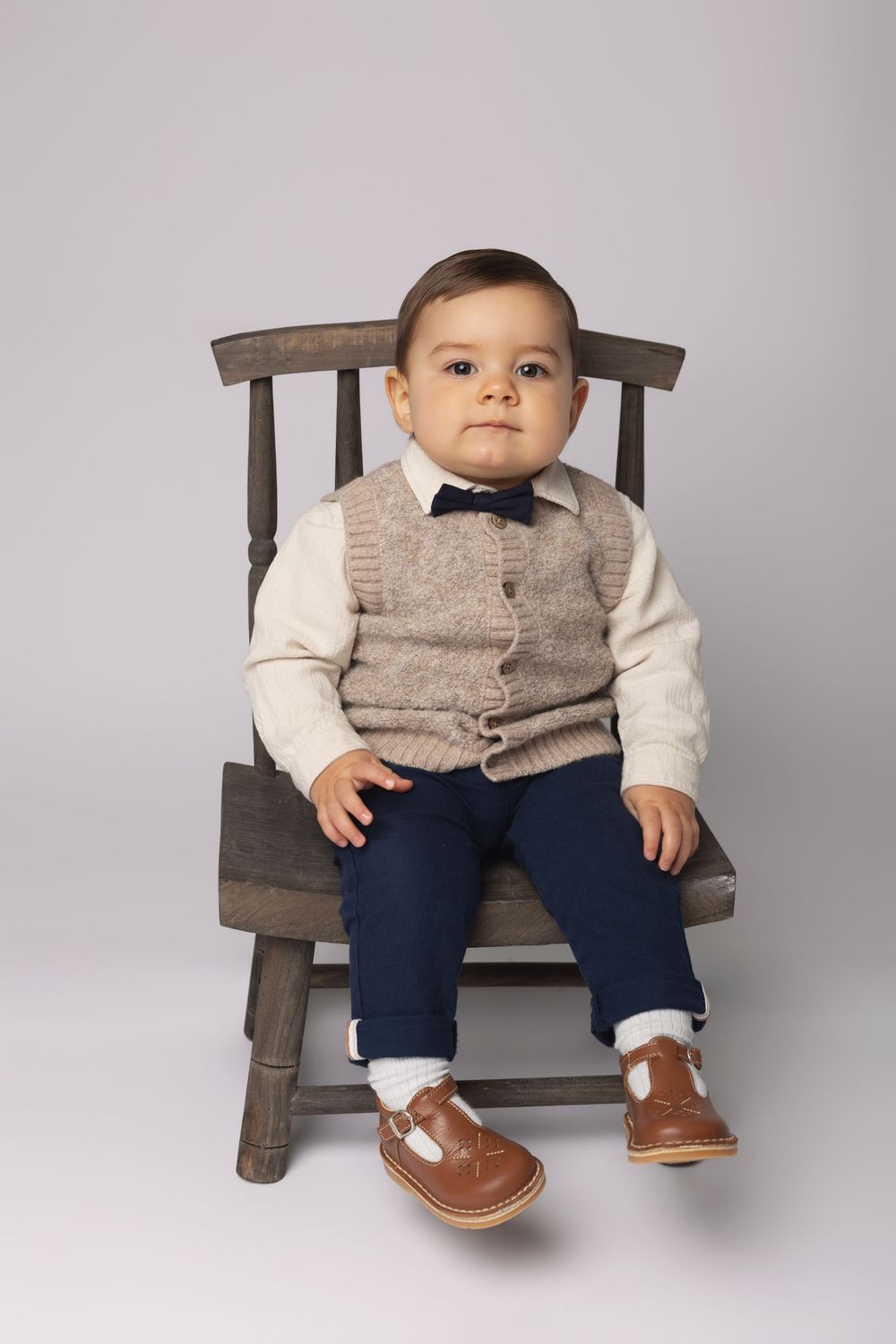 Adorable baby boy in bow tie, vest, navy pants, and brown shoes sitting on a wooden chair.