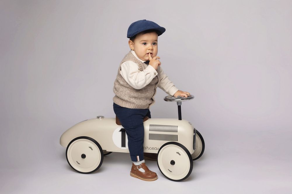 Toddler boy in navy cap and sweater vest sitting on a vintage cream ride-on toy car in a studio setting.