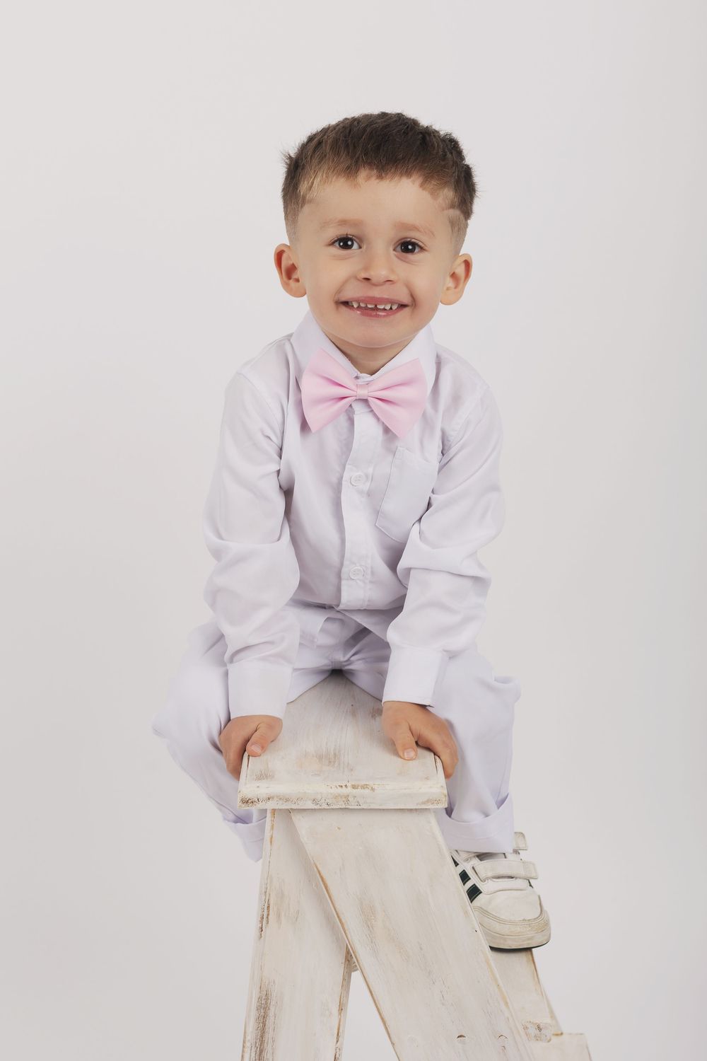 Young child in white dress shirt and pink bowtie poses with hands on white surface against light background.