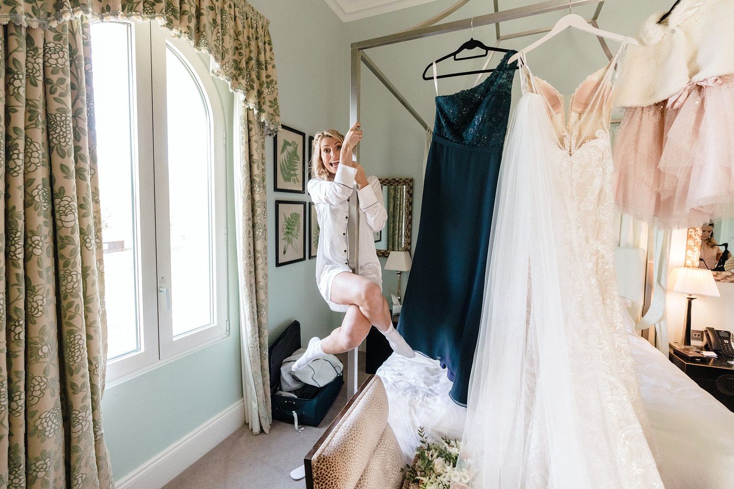 Wedding dress hangs from bed in a sunlit bridal suit at The Westin Nova Scotian room while bride hangs on bed.