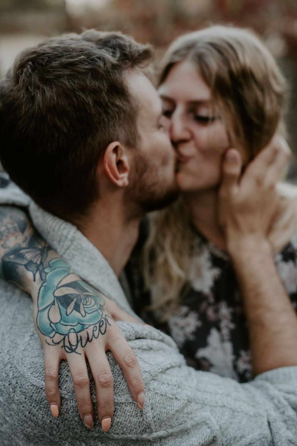 couple up close but slightly off focus kiss with  woman's tattooed hand in focus with the words "queen" in black in over blue flower