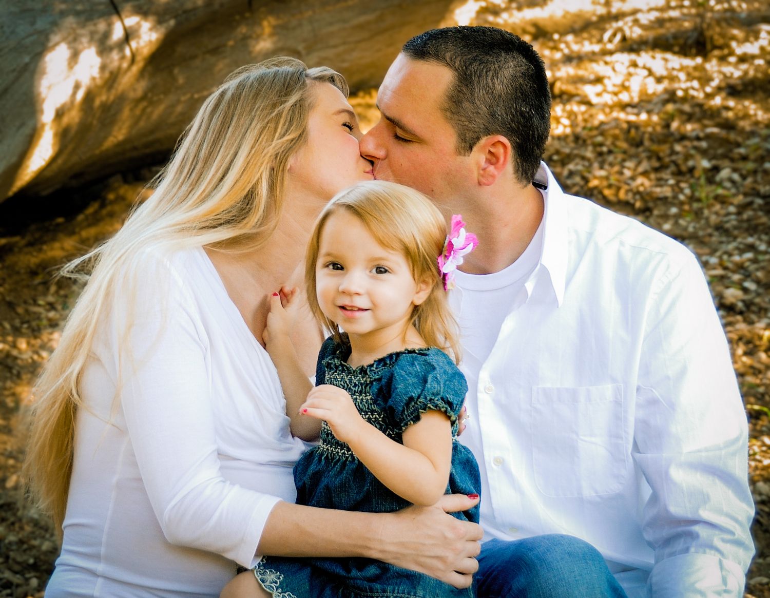 A family of three share an intimate moment outdoors against a golden-lit rocky background.