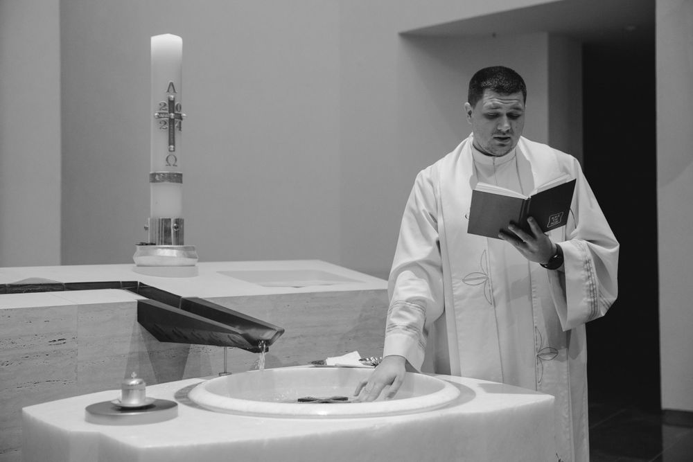A clergy member in white robes stands at a baptismal font while reading from a book.