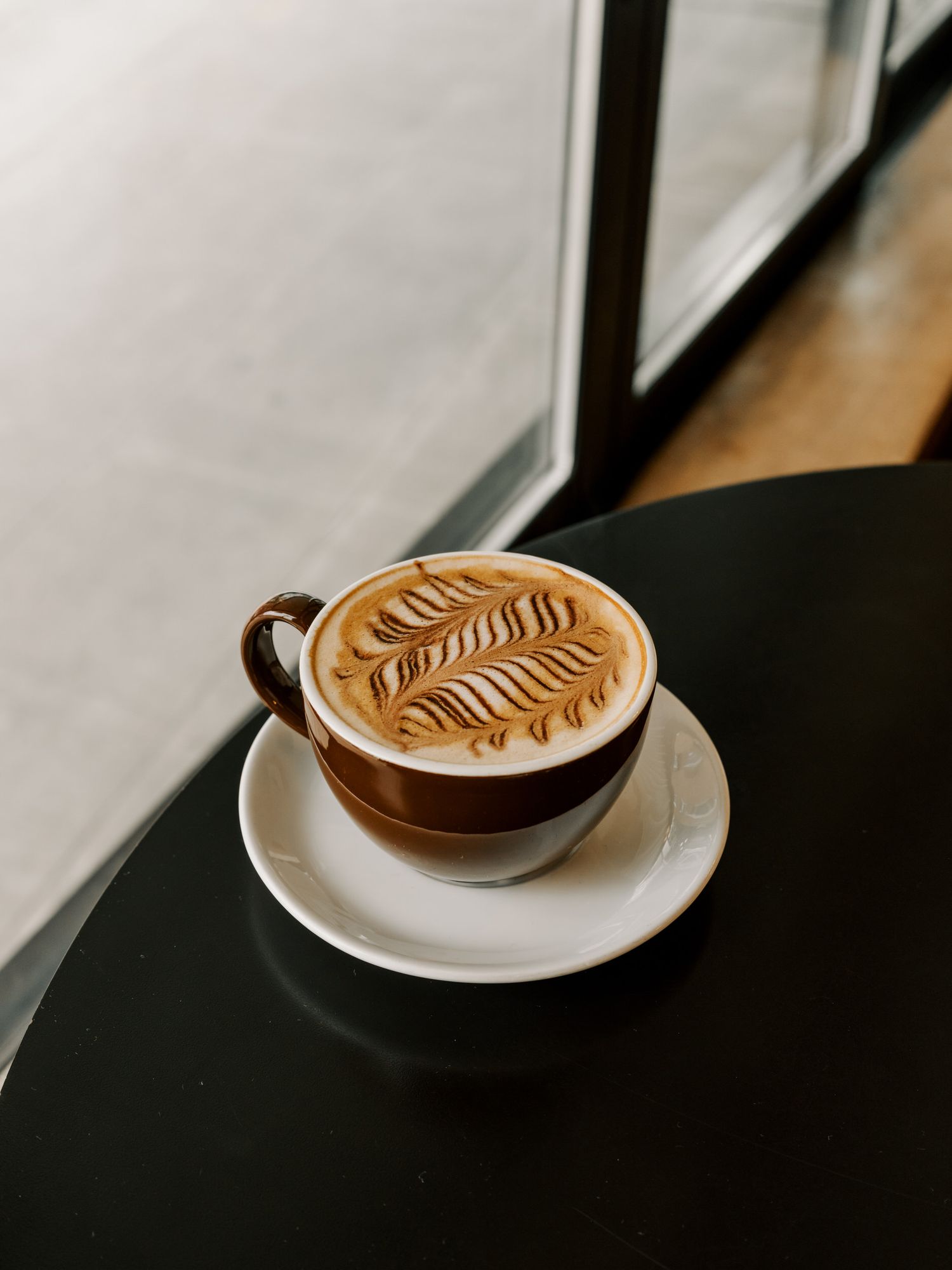 A white ceramic cup of latte with intricate fern leaf latte art pattern sits on a black table near a window.