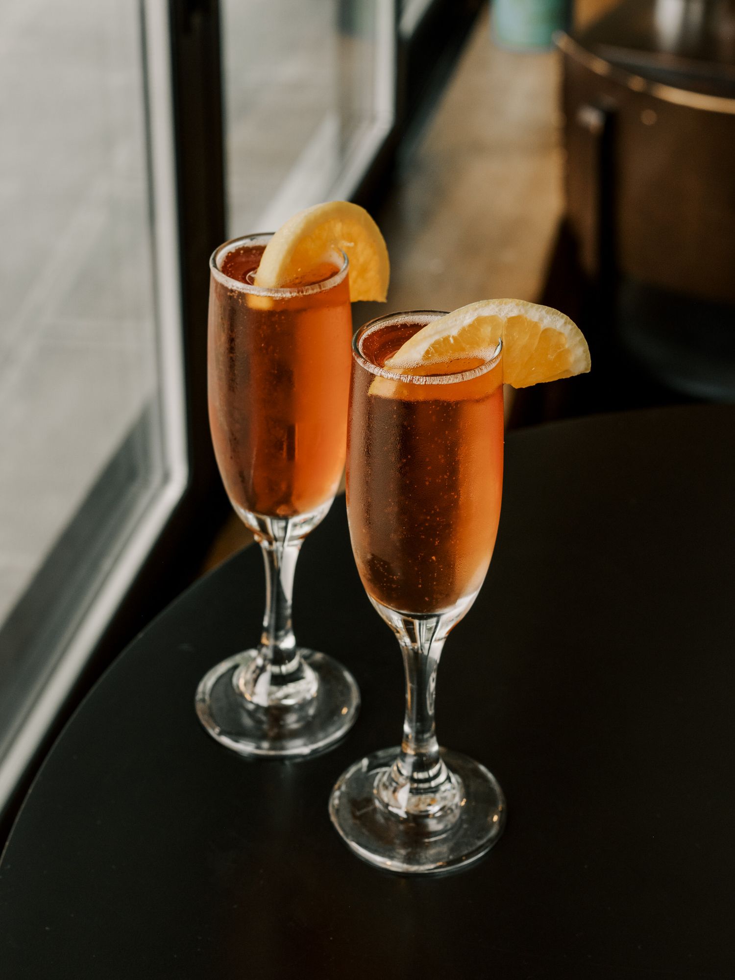 Two champagne flutes filled with orange-colored cocktails garnished with citrus slices sit by a window.