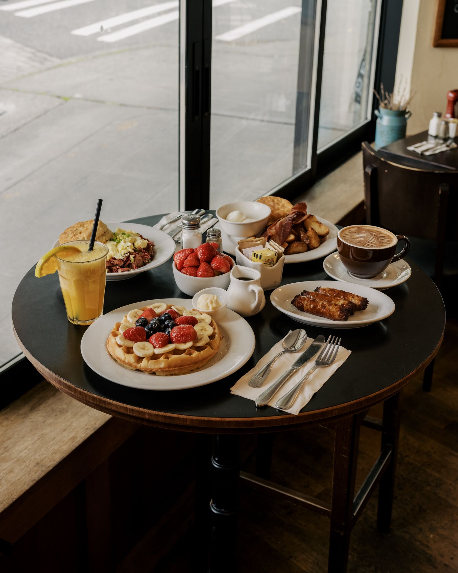 A breakfast spread on a round table featuring waffles with berries, coffee, orange juice, and pastries by a window.