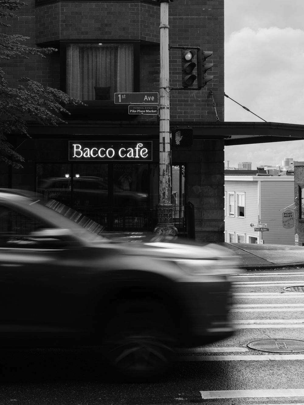 Black and white urban scene showing Bacco cafe storefront with motion-blurred car passing on wet street.