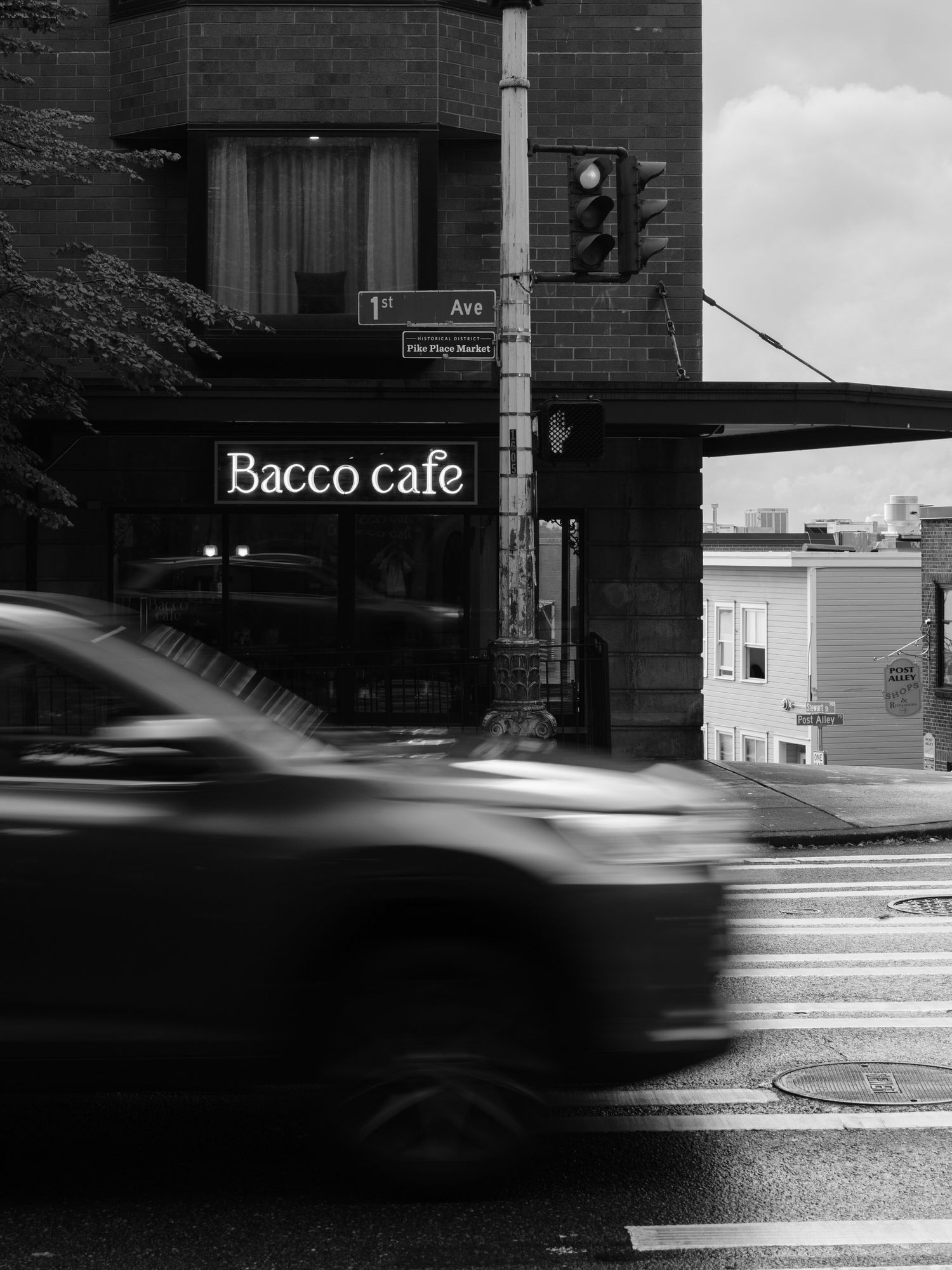 Black and white urban scene showing Bacco cafe storefront with motion-blurred car passing on wet street.