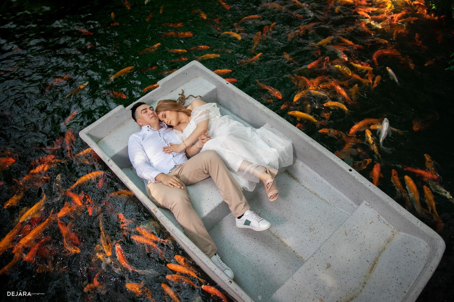 Creative wedding photo with couple lying in boat surrounded by orange koi fish.