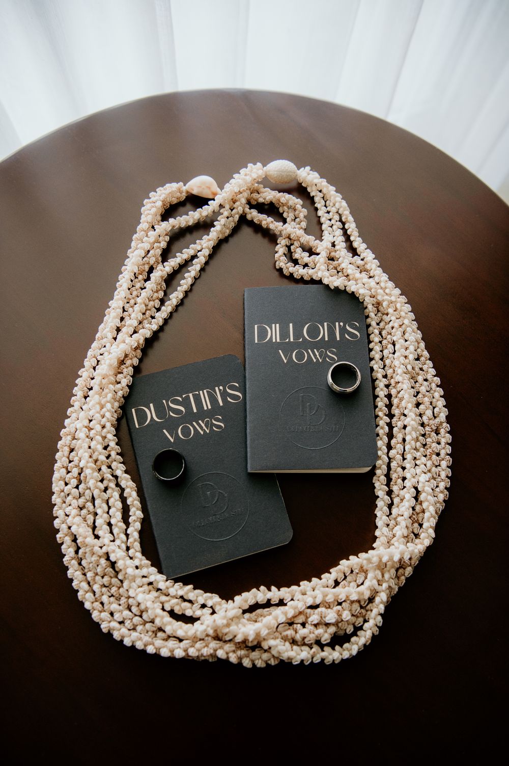 Two black vow books with wedding rings and pearl necklace displayed on dark wooden surface.
