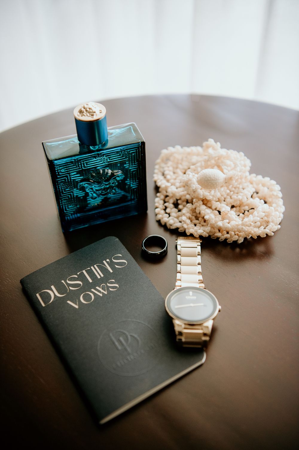 Wedding details including perfume, vows book, watch and pearl necklace arranged on dark table.