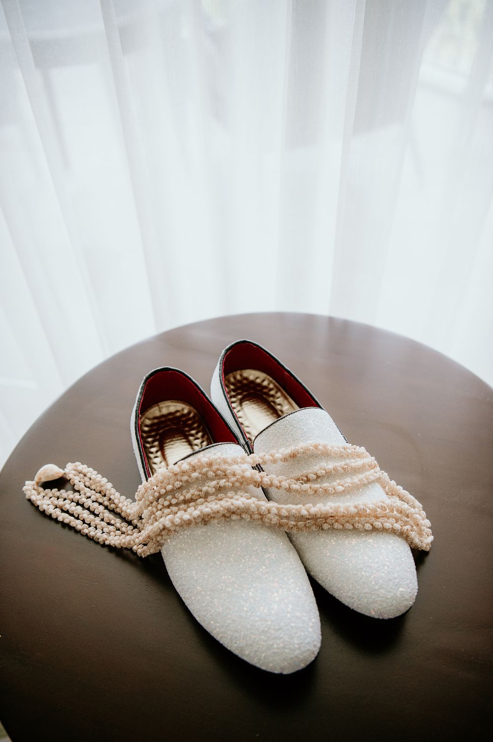 White glitter wedding shoes with pearl necklace on round brown table.