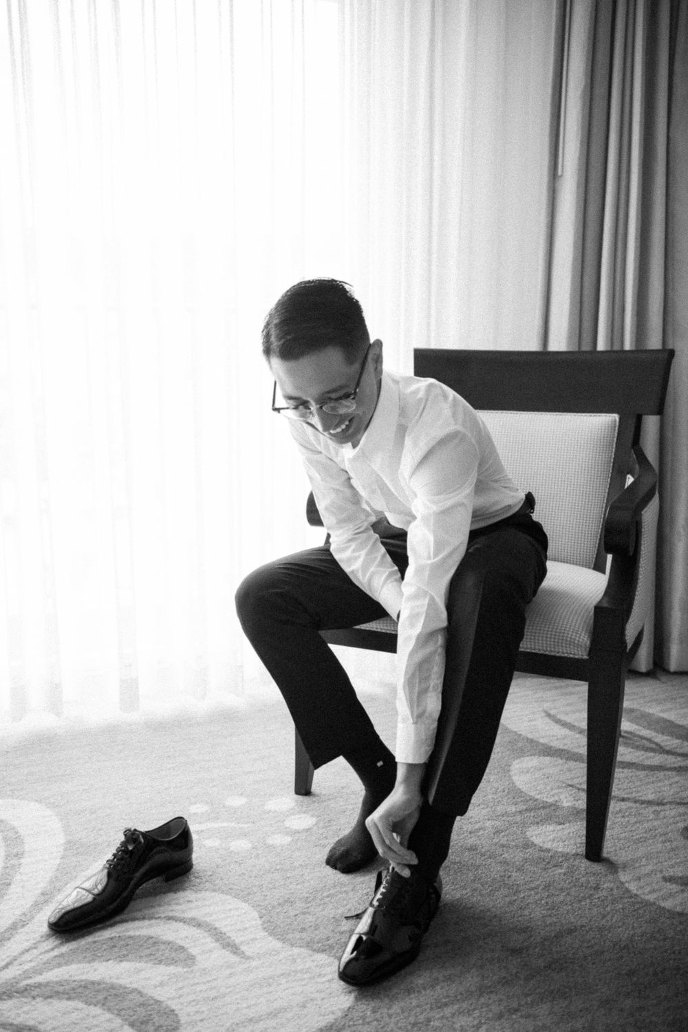 A person in a white dress shirt and glasses sits on a chair putting on formal black dress shoes.