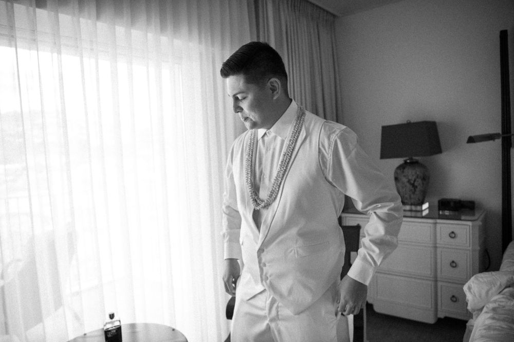 A person in an elegant white outfit stands by a window in a hotel room getting ready.