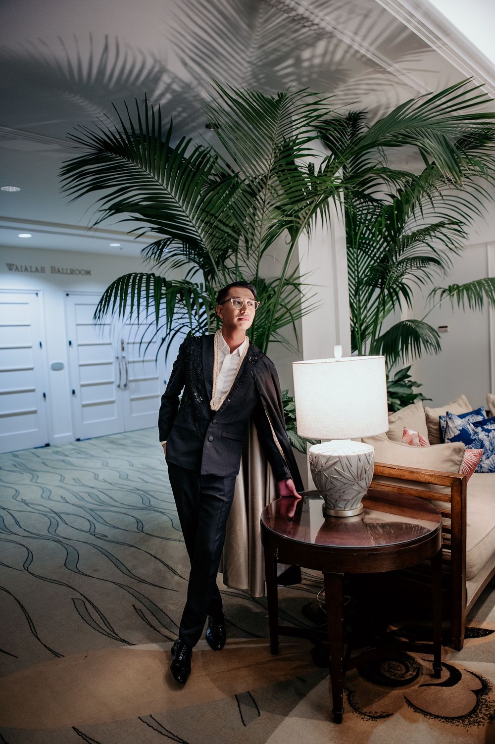 A person in a black suit poses against tropical palm plants in an elegant interior setting.