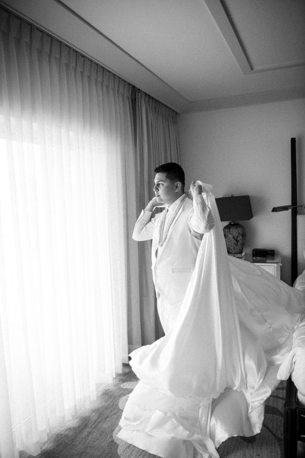 A person in white attire adjusts their outfit by a sunlit window in a hotel room.