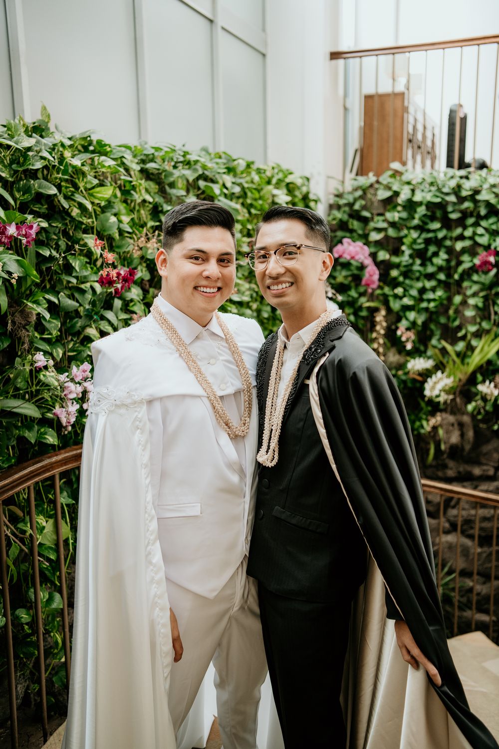 A couple in contrasting white and black formal attire pose together against a floral garden backdrop.