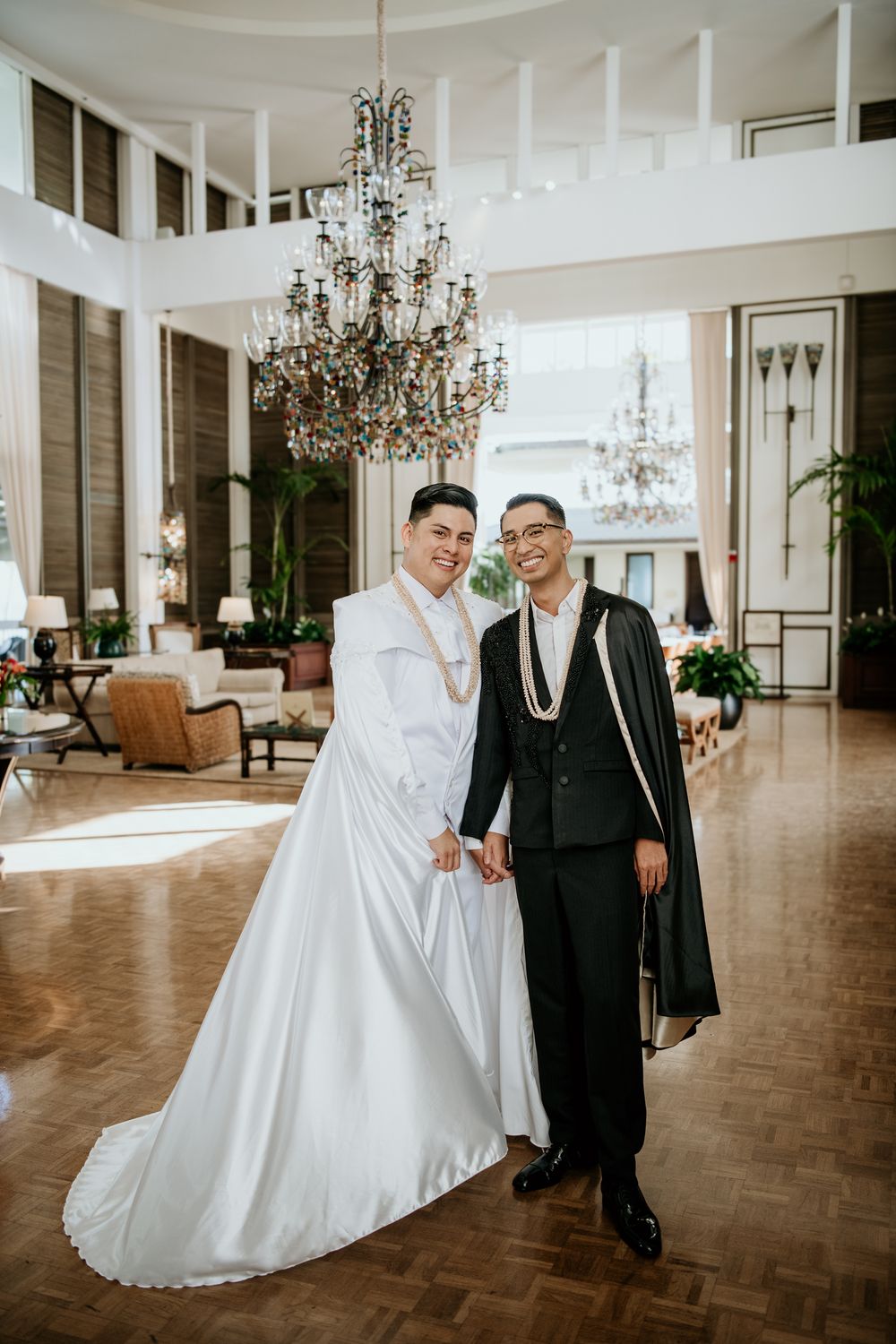 A wedding couple in coordinated white and black attire pose beneath an elegant crystal chandelier at the Kahala Hotel & Resort Lobby.