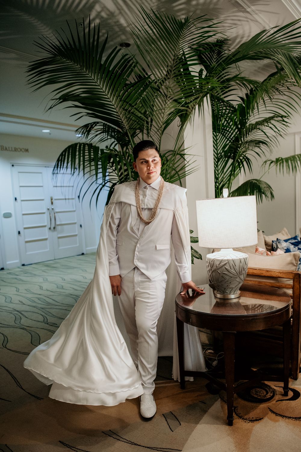 A person in an all-white formal suit and pearl necklace poses beside tropical palm plants.
