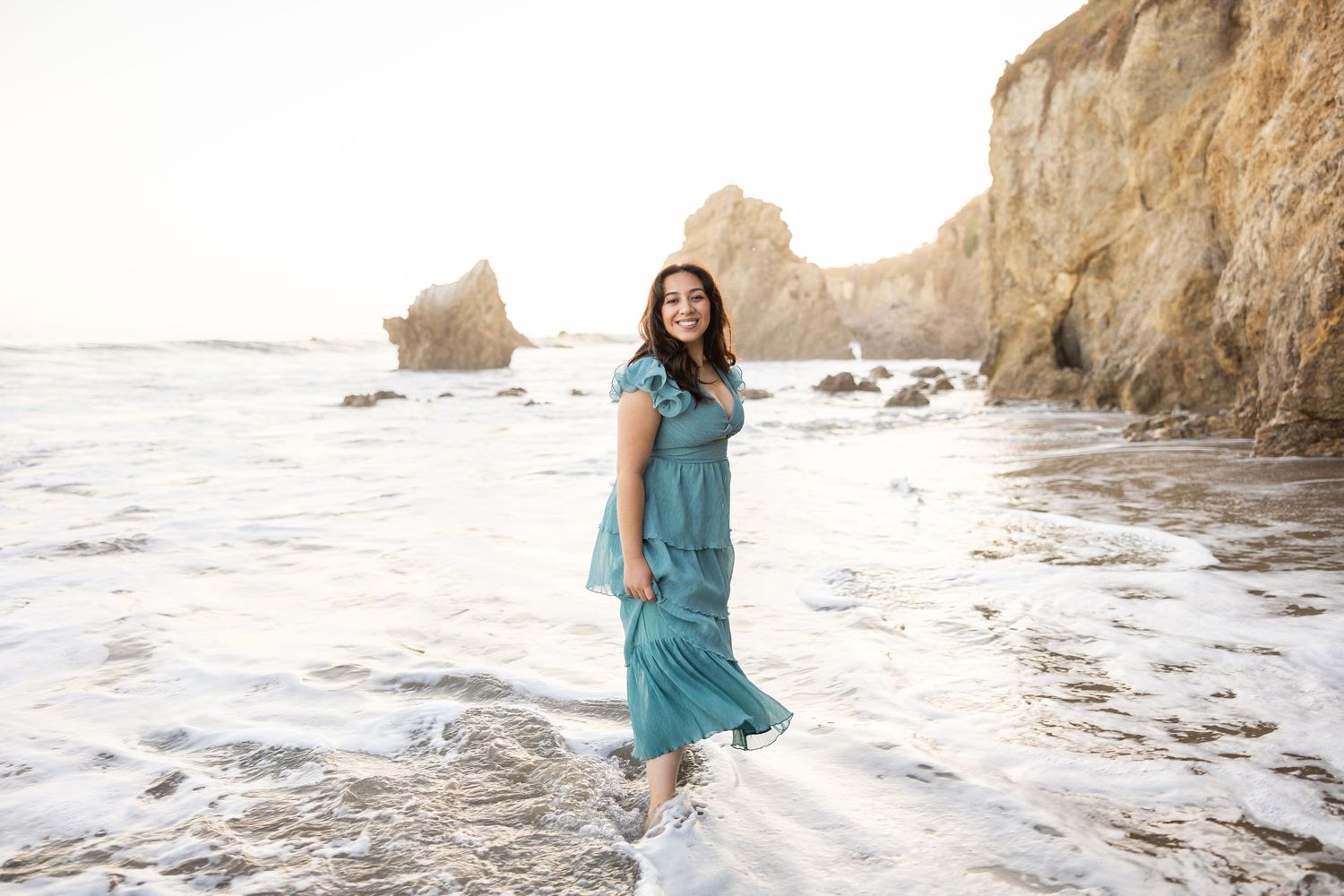 A person in a flowing turquoise dress walks along a rocky beach at sunset with coastal cliffs in the background.
