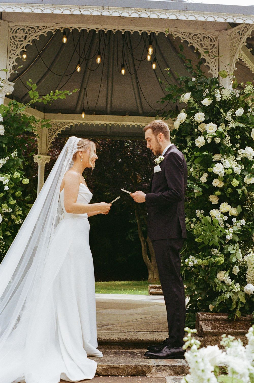 Couple exchanges vows during outdoor gazebo wedding ceremony surrounded by white roses and greenery.