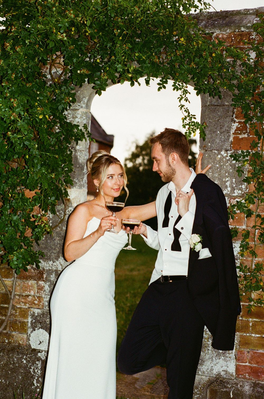 A couple in wedding attire sharing a toast under a garden arch with green vines and brick walls.
